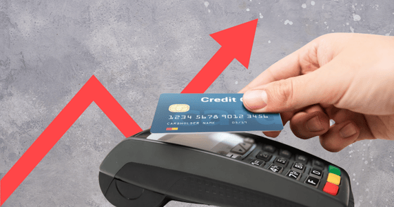 A hand taps a credit card to a payment machine in front of a rising red arrow against a gray background. 