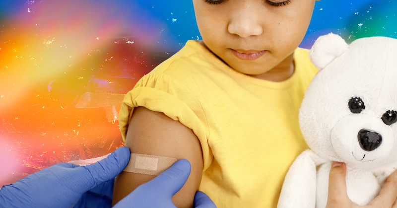 A young child holding a teddy bear receives a bandage on his arm against a rainbow background.