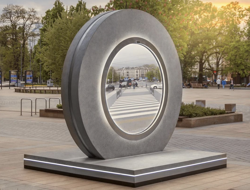 A circular sculpture in a city park with a screen in the center showing a different city streetscape.