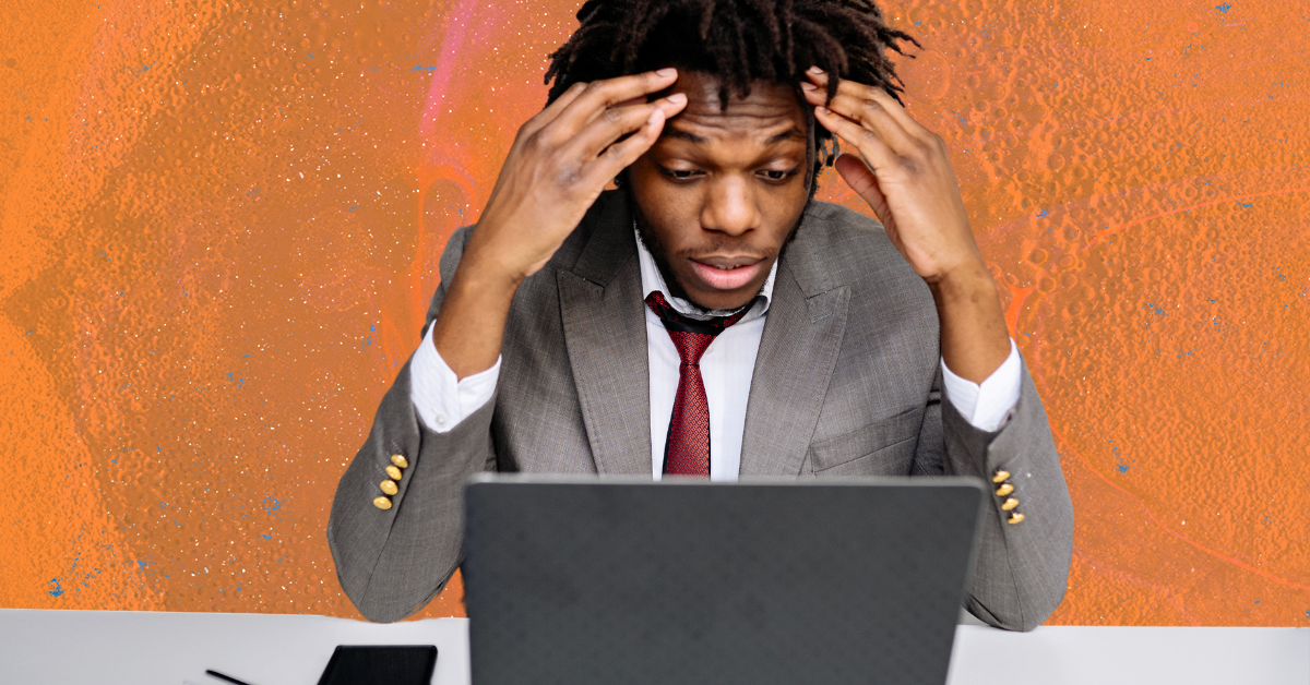 A black man in a gray suit and red tie holding his hands to his face and looking at a laptop screen on an orange background.